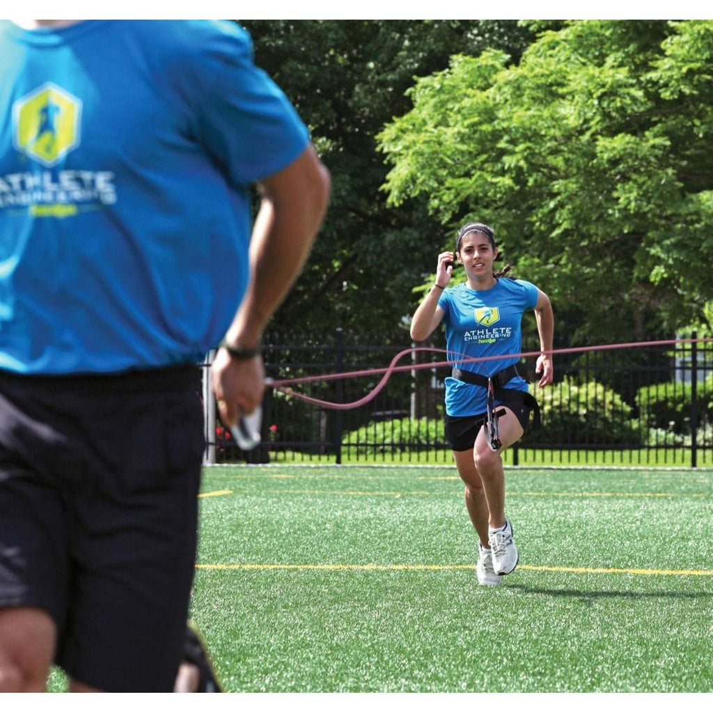 Person running on a sports field with another person in a blue shirt in the foreground.