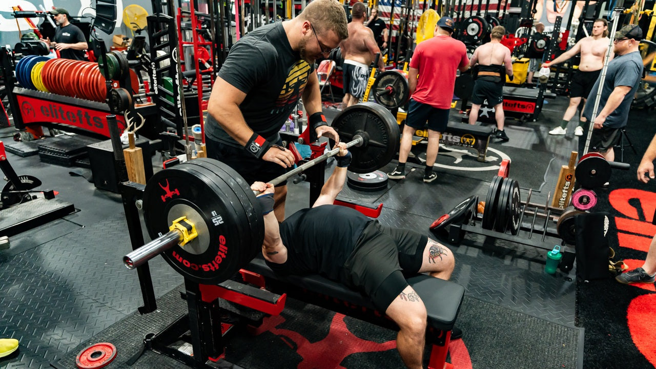 A lifter performs a barbell bench press with a spotter in the elitefts gym.